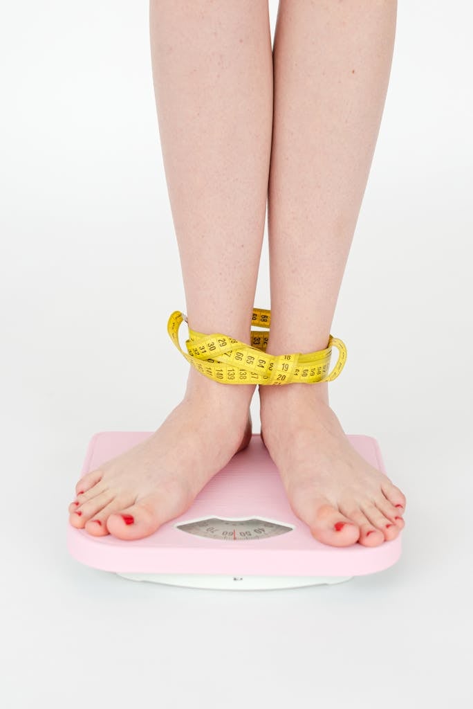 High angle of crop anonymous female with centimeter on legs checking weight on scales on white background